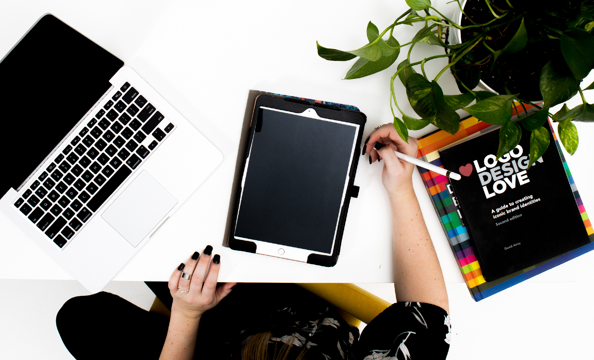 photo of a woman sitting at a white desk with her computer and tablet along with a logo design book and a plant